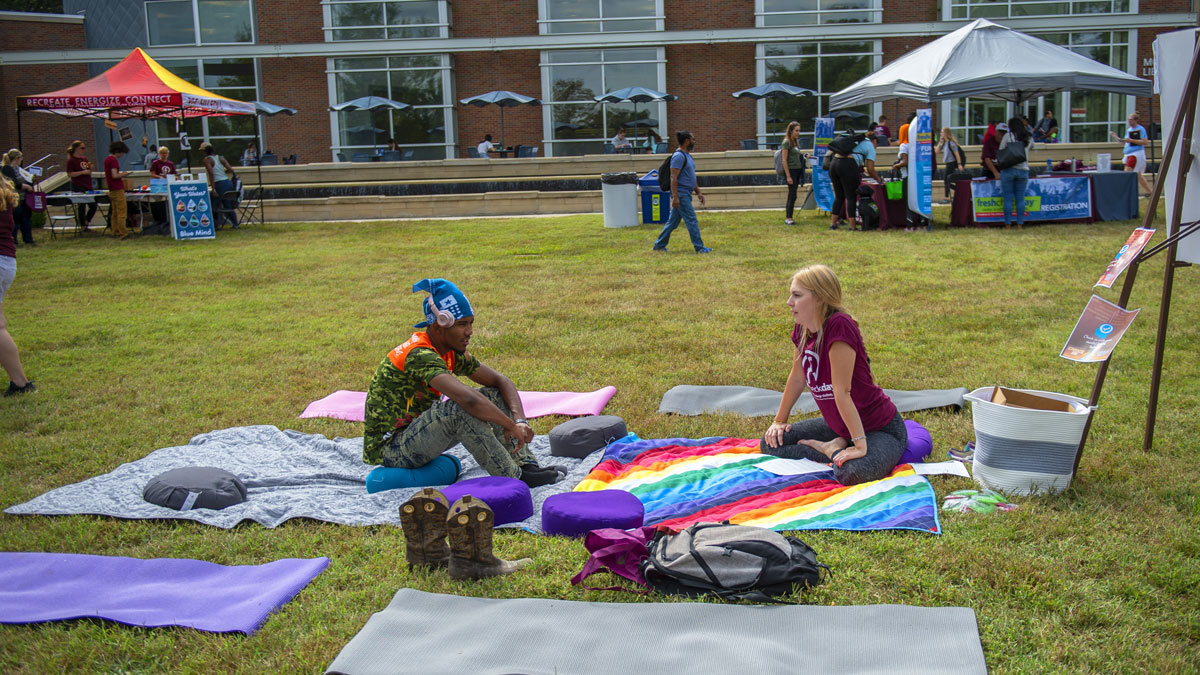 students at Fresh Check Day 2019, on the lawn at Morris Library.  A black man and white woman are seated on a blanket in a yoga/meditation area. Various tents are visible in the background.