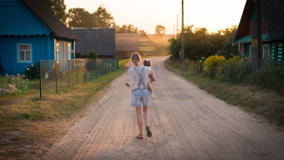 woman walking down a dirt road with houses on either side, holding a small child