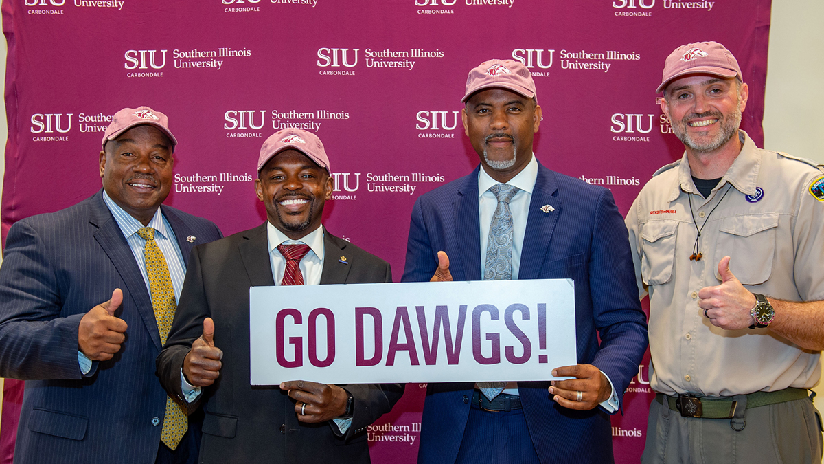 Four men, three black and one white, smiling at the camera. They are holding a sign that says 'Go Dawgs'