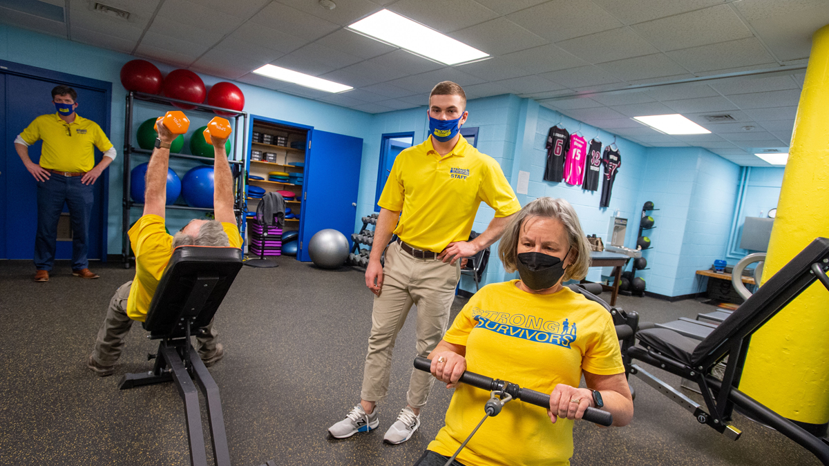 A woman, right, exercises with assistance from a senior exercise science major. At left, a man, participates in workouts with her, as Strong Survivors director, watches.