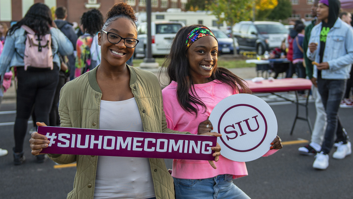Two young black women are smiling while holding signs. One sign says #SIUHOMECOMING, and the other sign says SIU. There are people seen in the background, walking around.