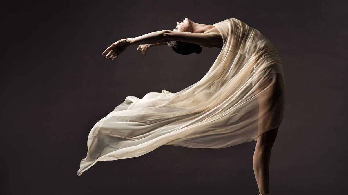 Image of a female dancer, draped in a gauzy fabric. She is bent slightly backward, giving the illusion that the wind is blowing her over.