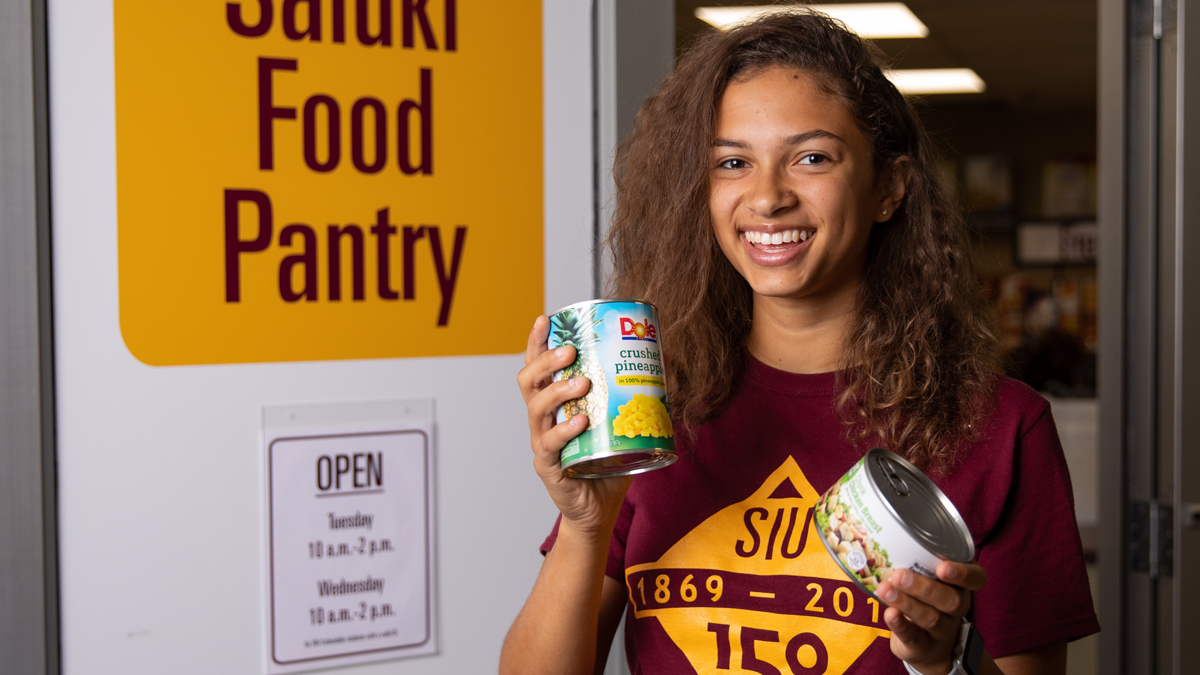 Young black woman, smiling, holding a can of crushed pineapple in one hand and canned chicken in the other.