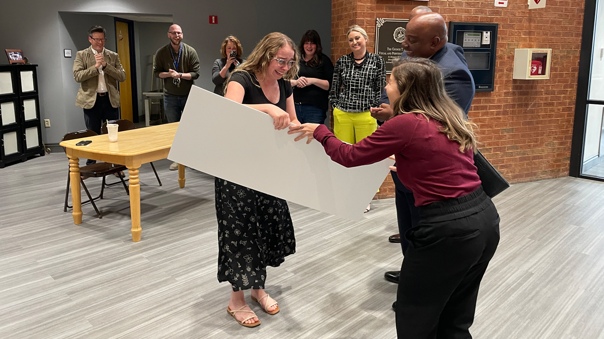 A young woman is receiving an award. She is a white woman with blonde hair and glasses. She is wearing a black shirt and a black and white skirt, with sandals. She is in a room with several others who are smiling and applauding. She is smiling and crying while accepting her award.