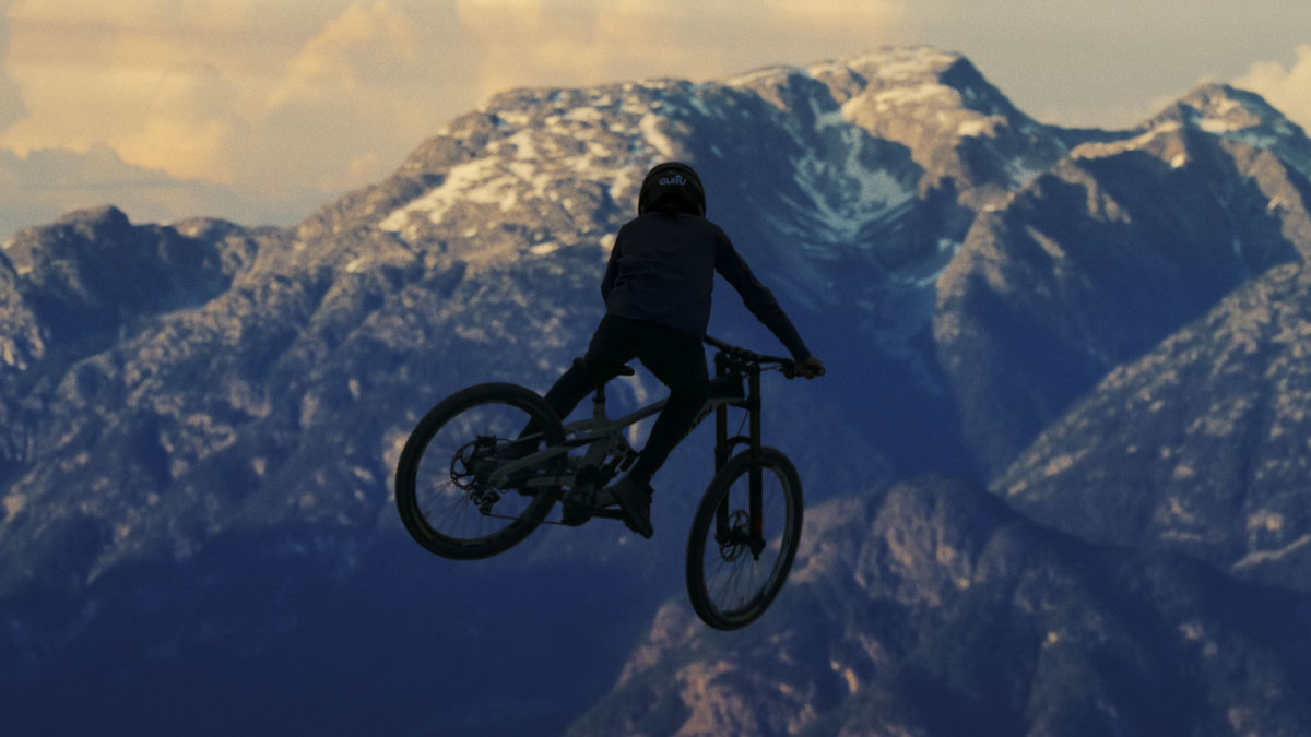 Image of a person in midair, riding a mountain bike. A mountain range is visible in the background.