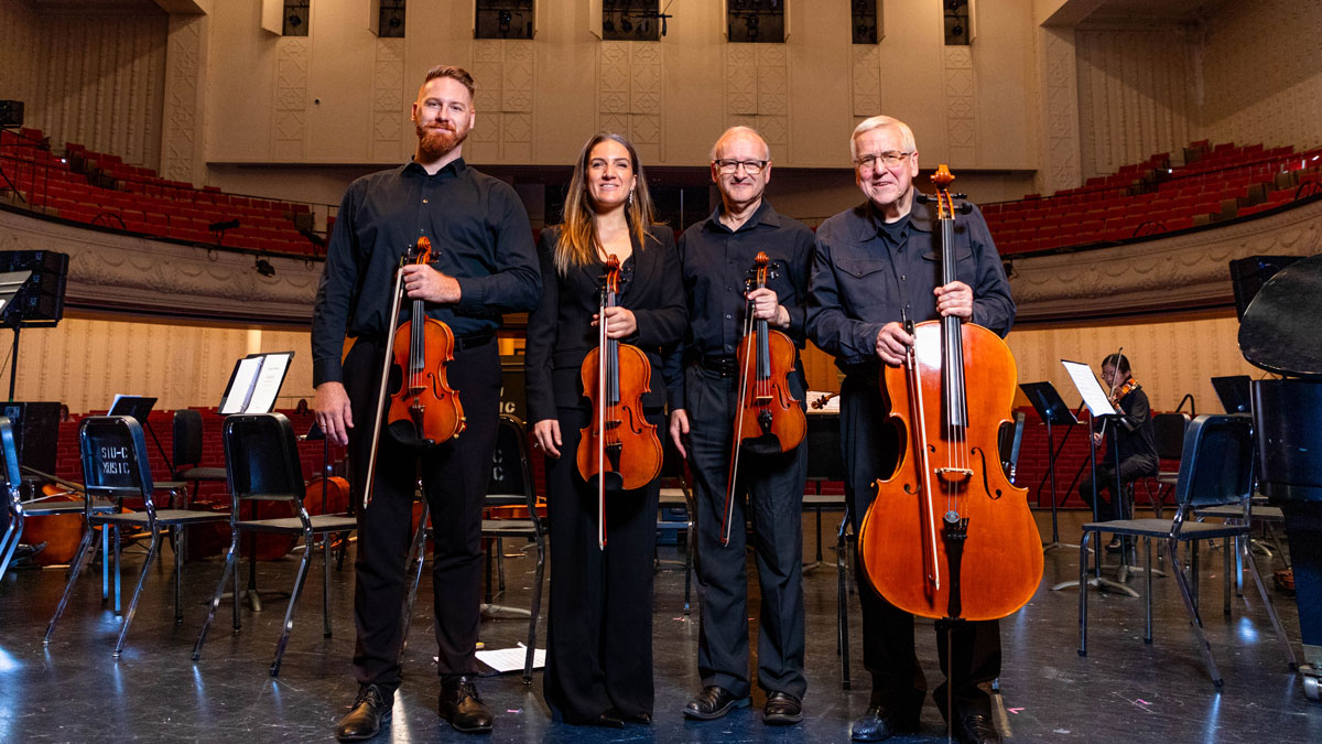 Four musicians stand on a stage, with their string instruments