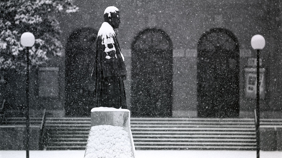 Delyte Morris statue on the campus of Southern Illinois University. Shryock Auditorium is seen in the background. It is snowing. The steps, trees, and statue are all covered in snow.