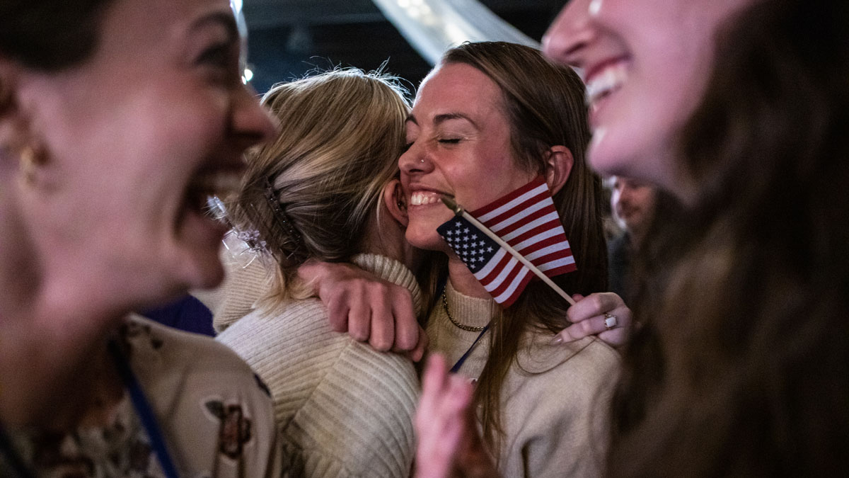 Two women are embracing. One is smiling and one is holding a small American flag. In the foreground you can see two other women, laughing and smiling.