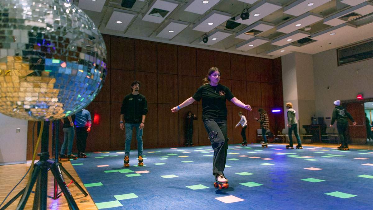 Students are seen skating in a large room converted to a skating rink. There is a disco ball in the foreground.