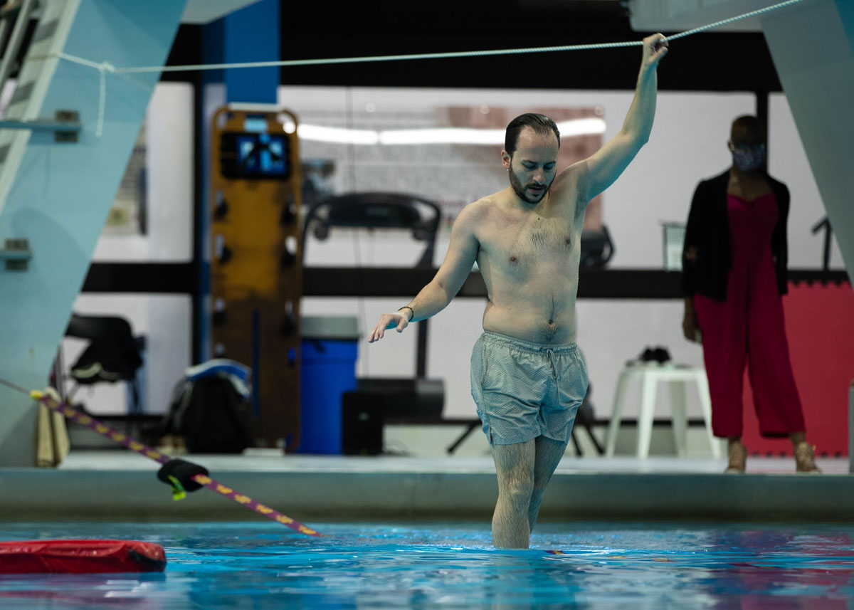 A man is seen in swim trunks and no shirt. He is walking on a slack line, over a pool, while holding another line above his head.