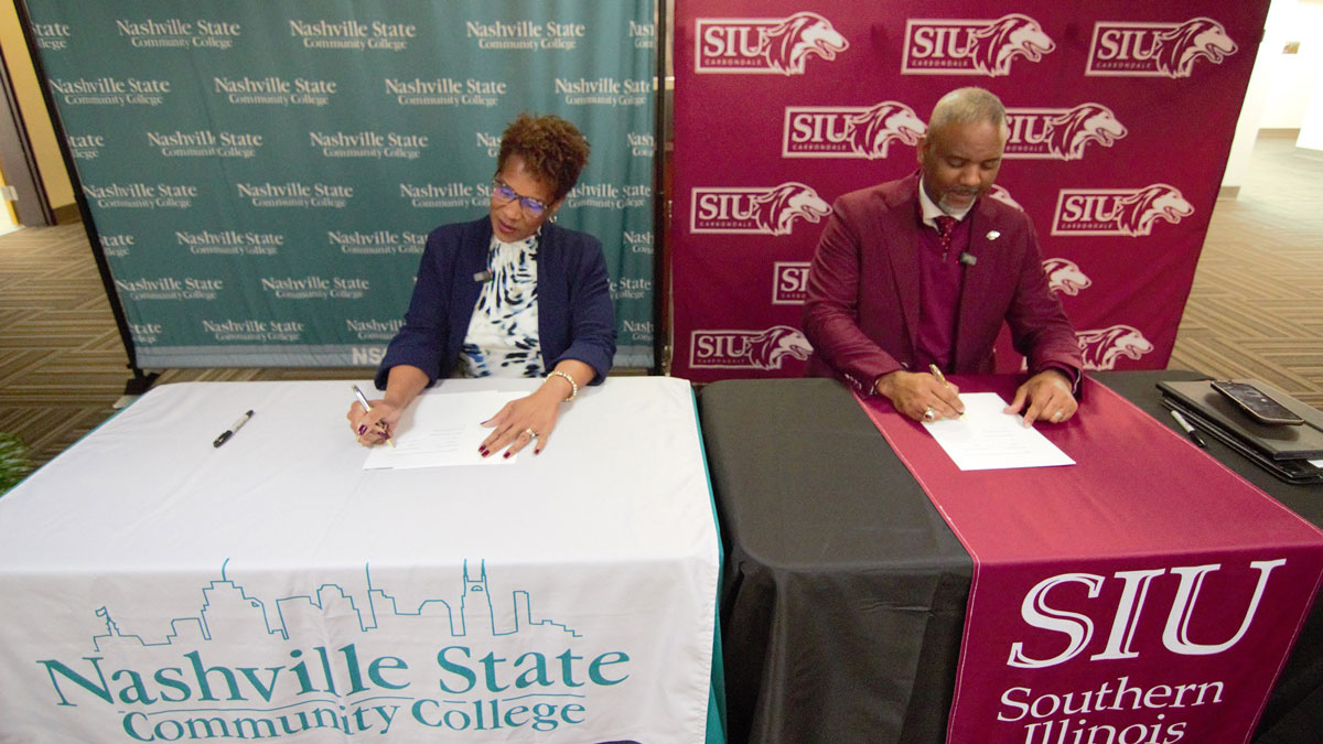 A black woman and a black man are seen seated at individual tables. Each table is draped in insignia from their respective schools. Both are looking down at the papers before them as they sign.