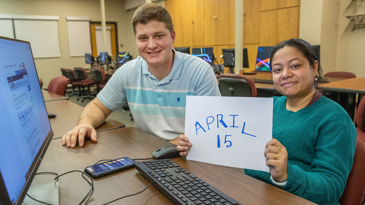 A man and woman are seated in front of a computer. She is holding a sign that reads April 15.