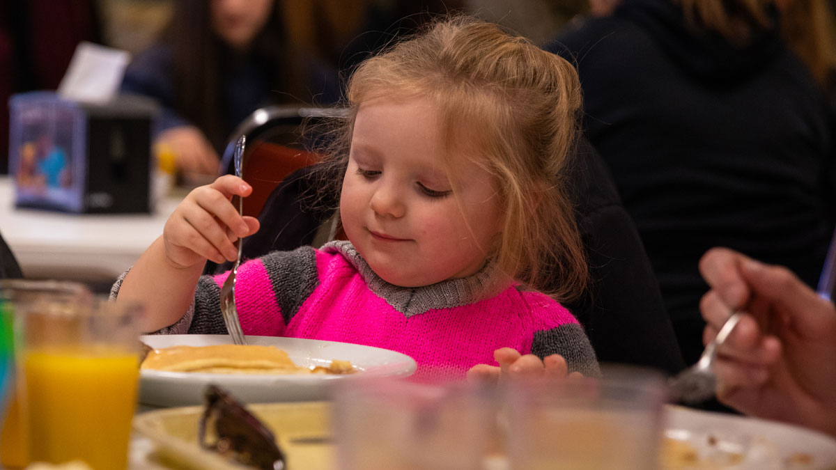 Young blonde girl is seated at a table, eating pancakes.