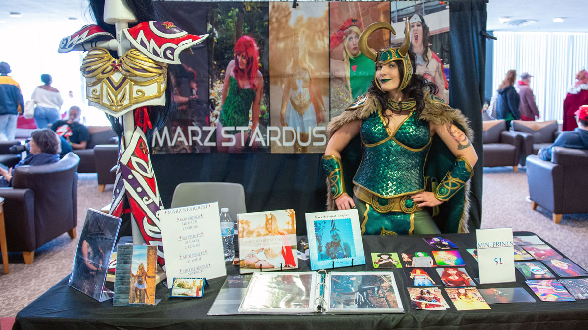 A woman is dressed in a green cosplay costume while working a vendor booth.