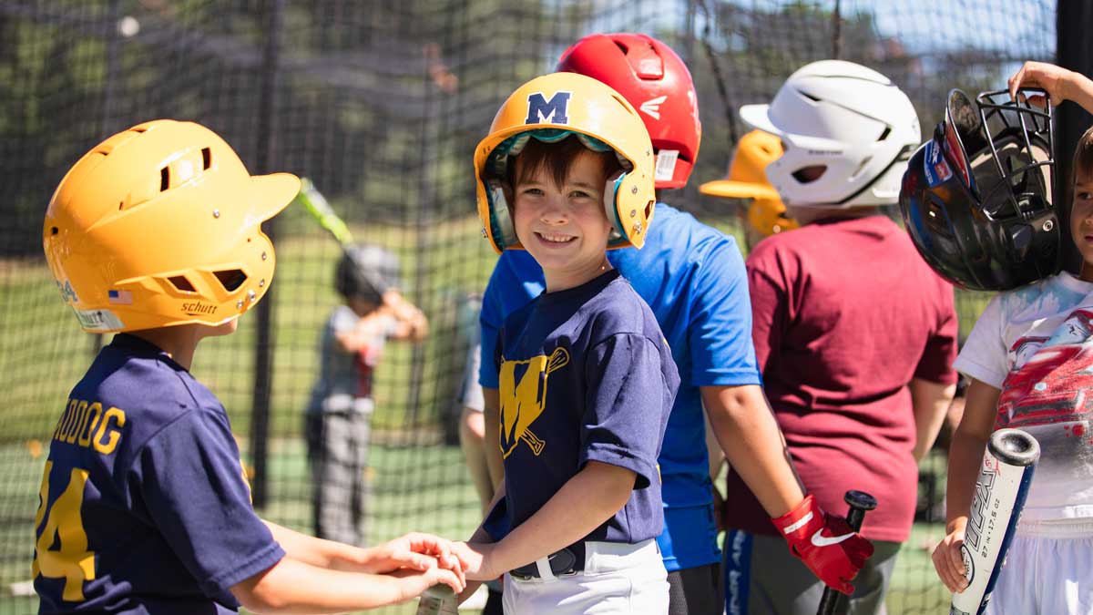 Children are waiting their turn to play baseball. One young man is half-turned, smiling at the camera. He is wearing a blue and yellow baseball jersey and cap.