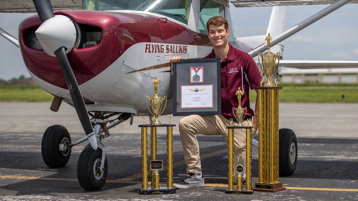  A young man is kneeling on a tarmac, beside an airplane. He is surrounded by trophies, and is holding a framed medal and certificate.
