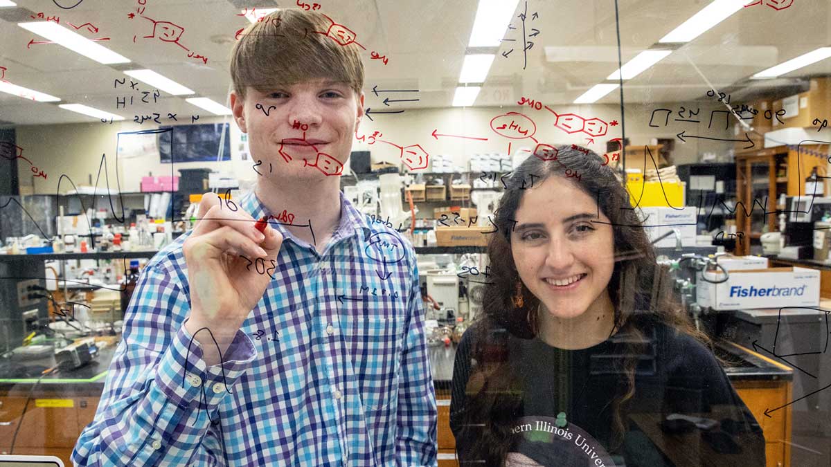 A young man and woman are seen through plexiglass. There are equations written all over the surface.