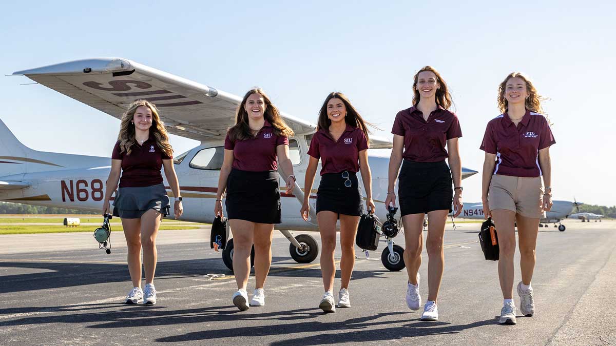 Five young women are walking toward the camera. Small airplanes are visible in the background.