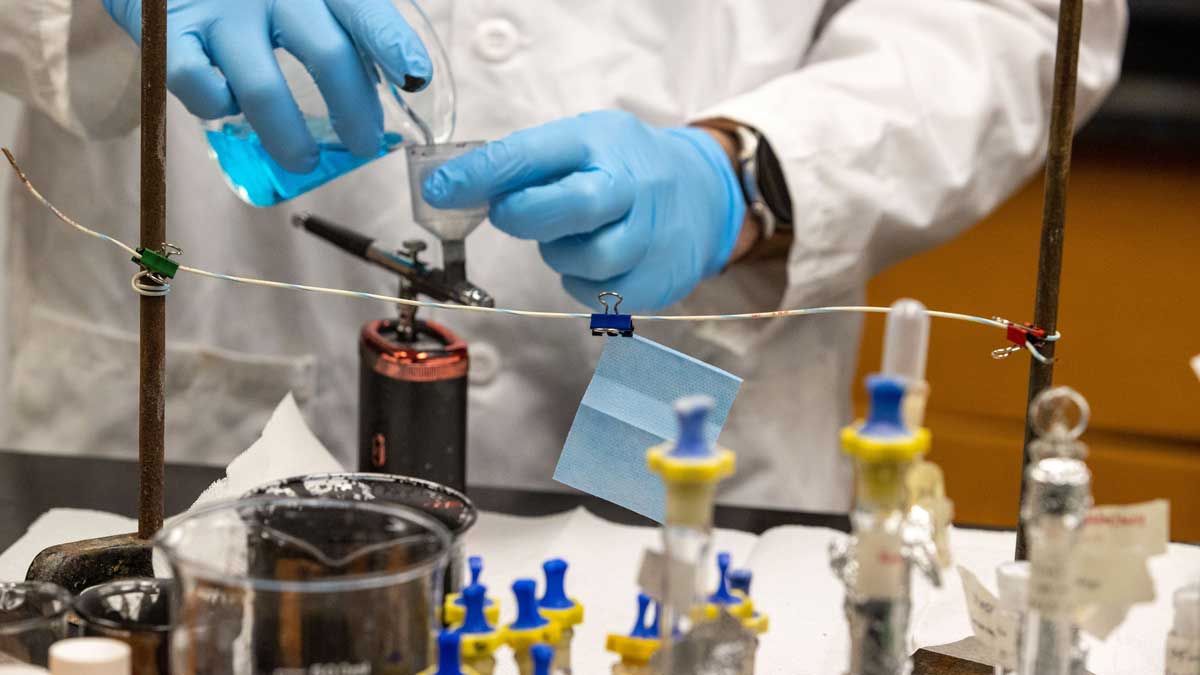 Image of what appears to be a lab. A person is wearing surgical gloves while pouring liquid into a glass container