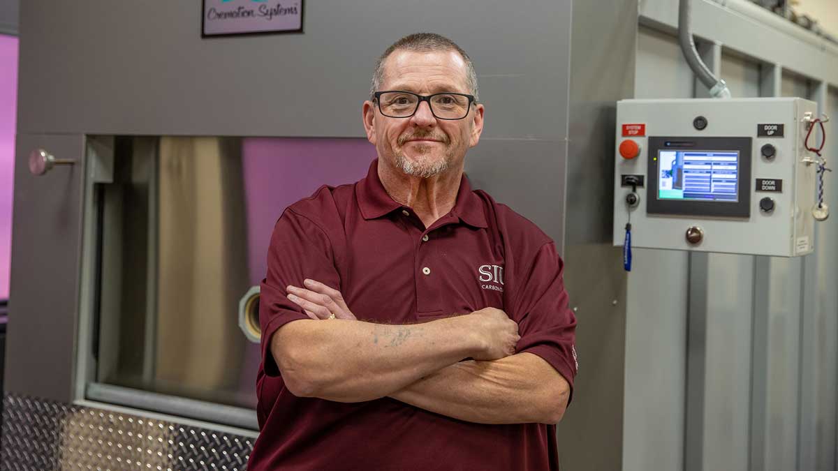 A man wearing a maroon shirt, with arms crossed over his chest, is standing in front of a crematory.