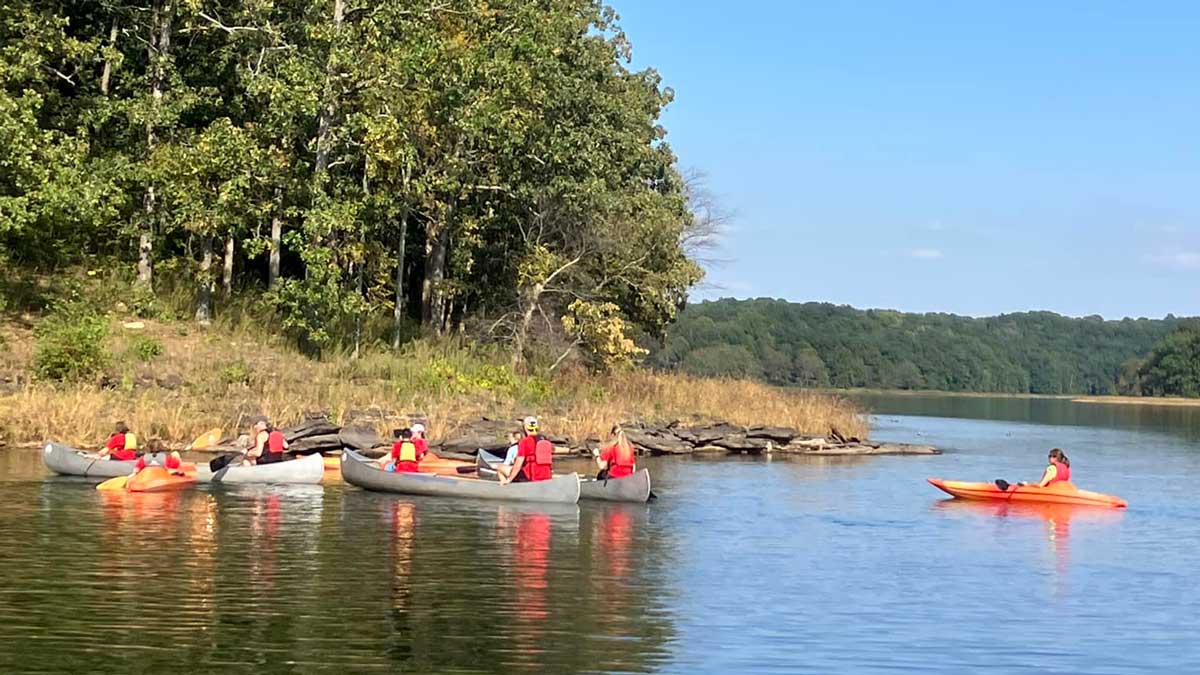 Image of people in canoes and kayaks. They are on the water and trees and rocks are visible in the background.