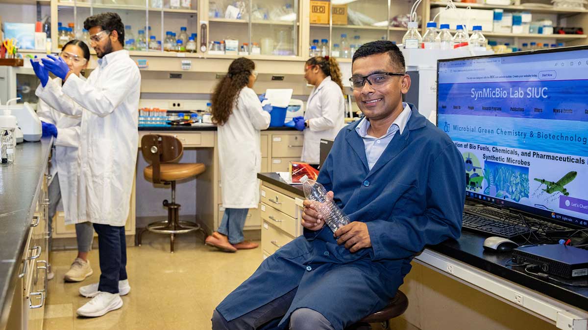  A man is seated in a lab, smiling while looking at the camera. In the background are others, wearing lab coats, and talking.