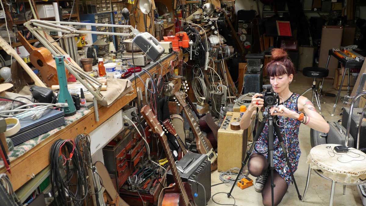 A woman is setting up a camera on a tripod. She is in a room filled with musical instruments, cords, cameras, and lights.