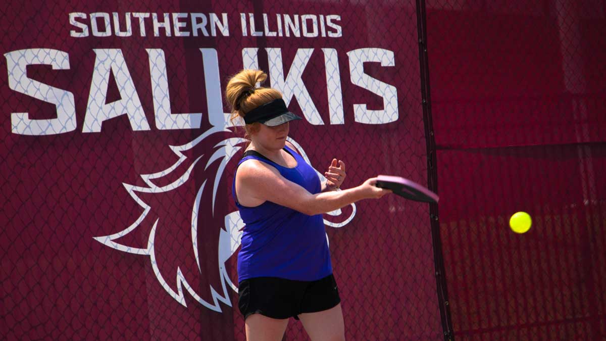 Image of a woman playing pickleball. Behind her is a fence covered in maroon. It says Southern Illinois Salukis,