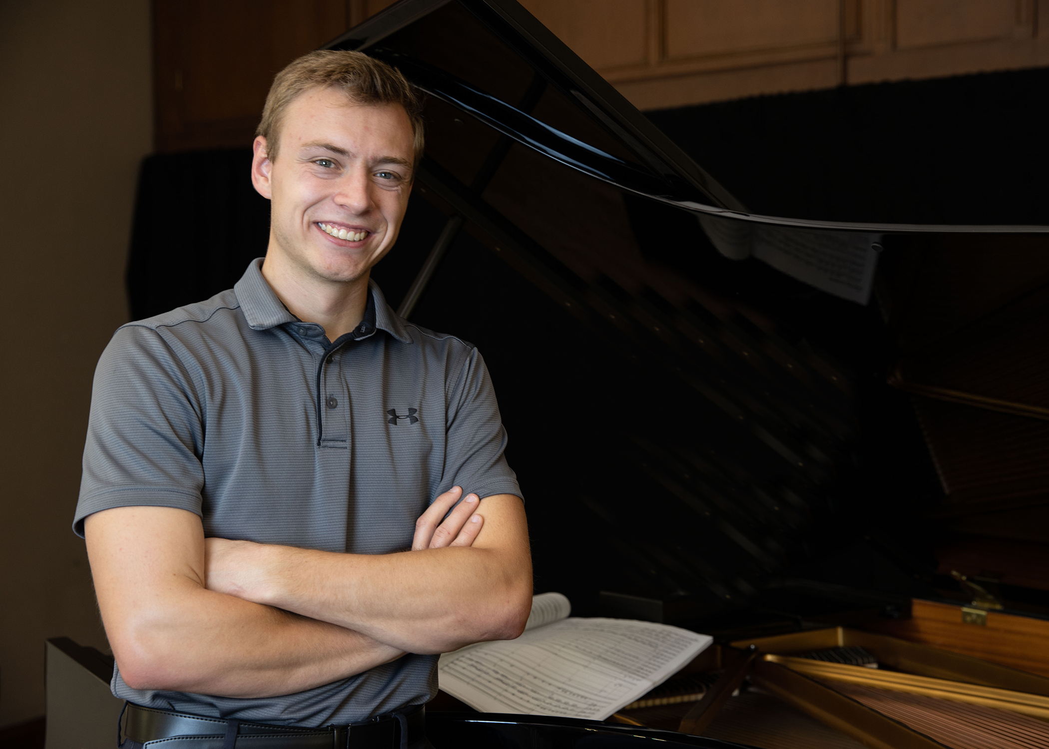 A young man wearing a grey short sleeved shirt, with a collar, is smiling. His arms are crossed as he leans on a piano. Sheet music is visible in the background.