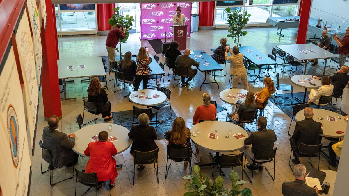 A group of people, seated at tables, are listening to a speaker.