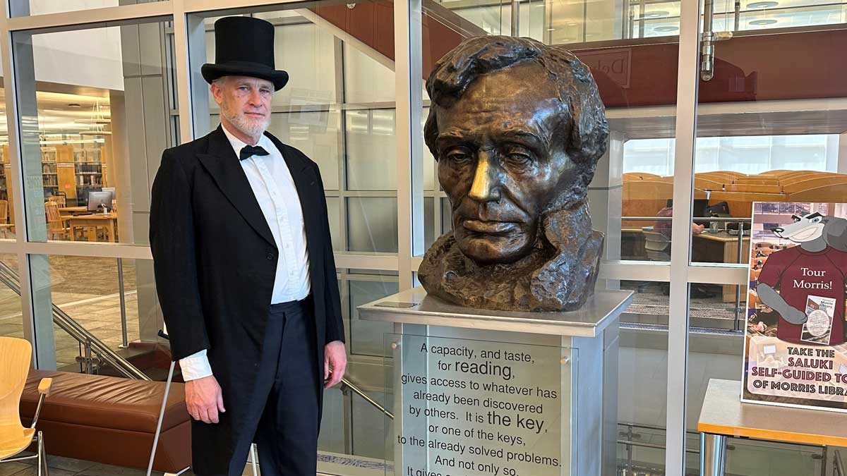 A man is standing next to a bronze bust of Abraham Lincoln. The man is dressed in a top hat and black jacket with tails.