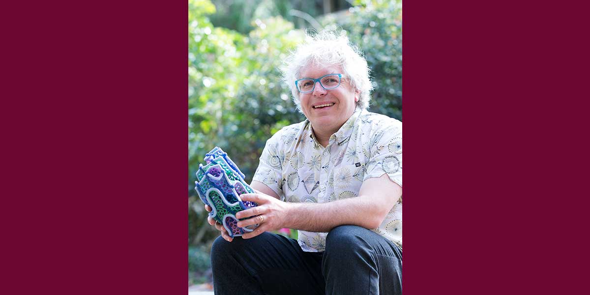 A man with white hair is smiling. He is wearing a short sleeve shirt, blue jeans, and blue glasses. He is holding a gyroid.