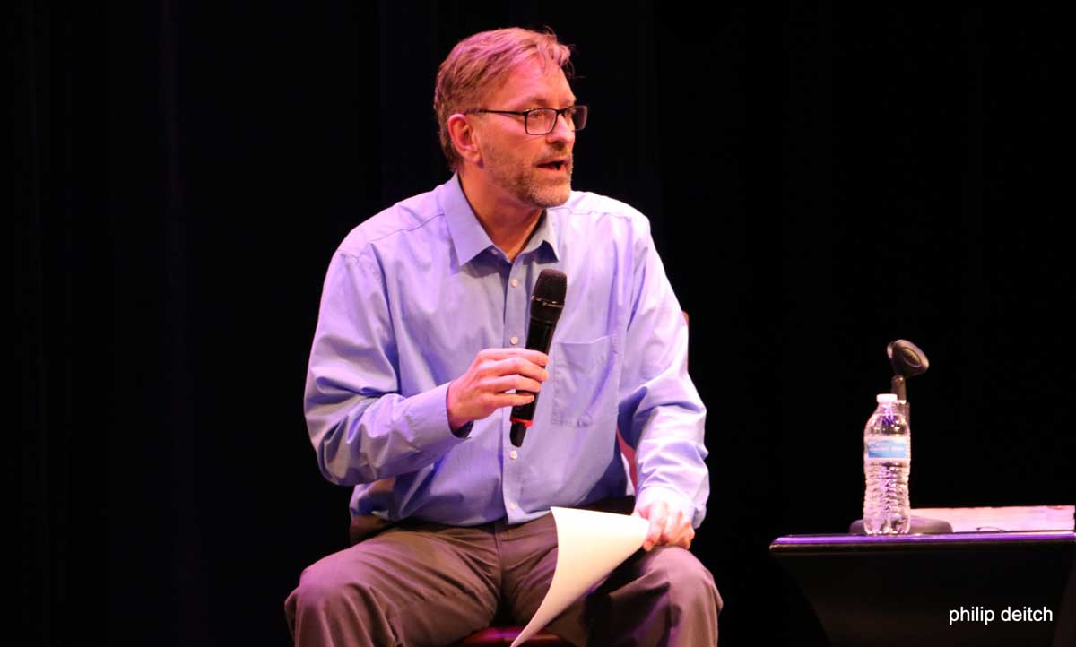  A man is seated in a chair, on a stage, talking