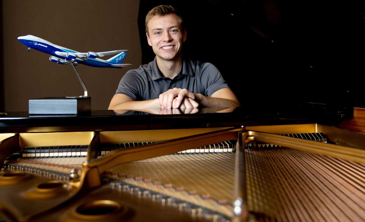 A young man is seated at a piano. The strings inside are visible. There is also a small statue of an airplane in flight