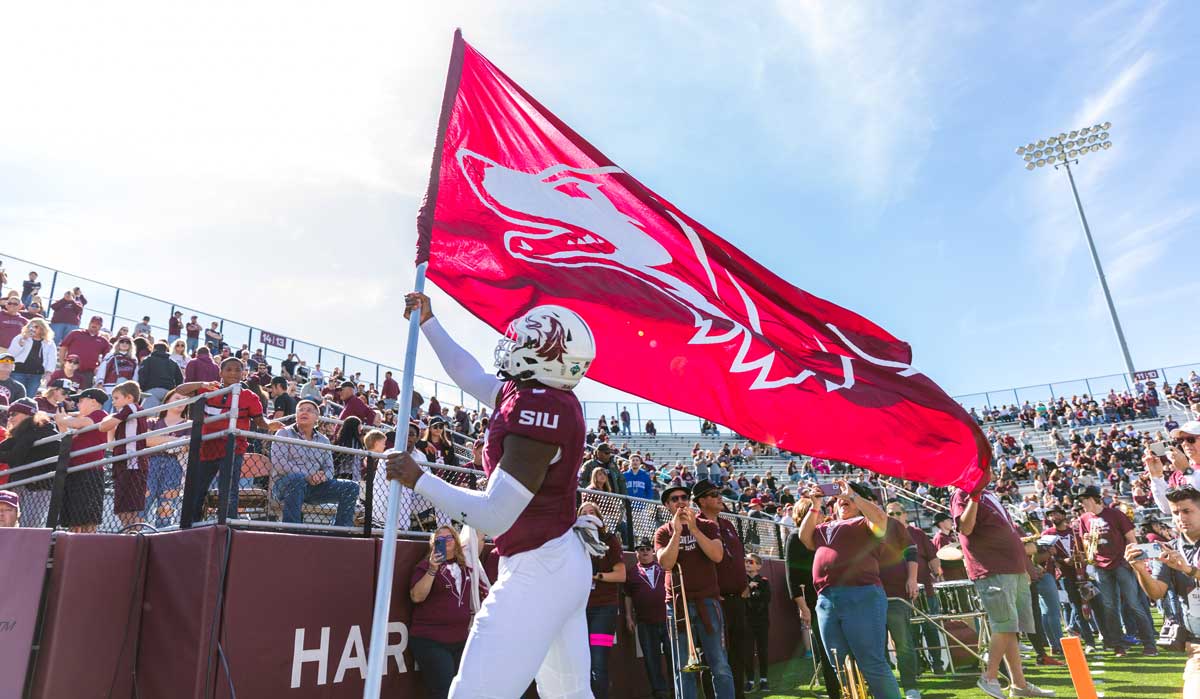 A football player is waving a flag with the Saluki Athletics logo, in front of a crowd of spectators.