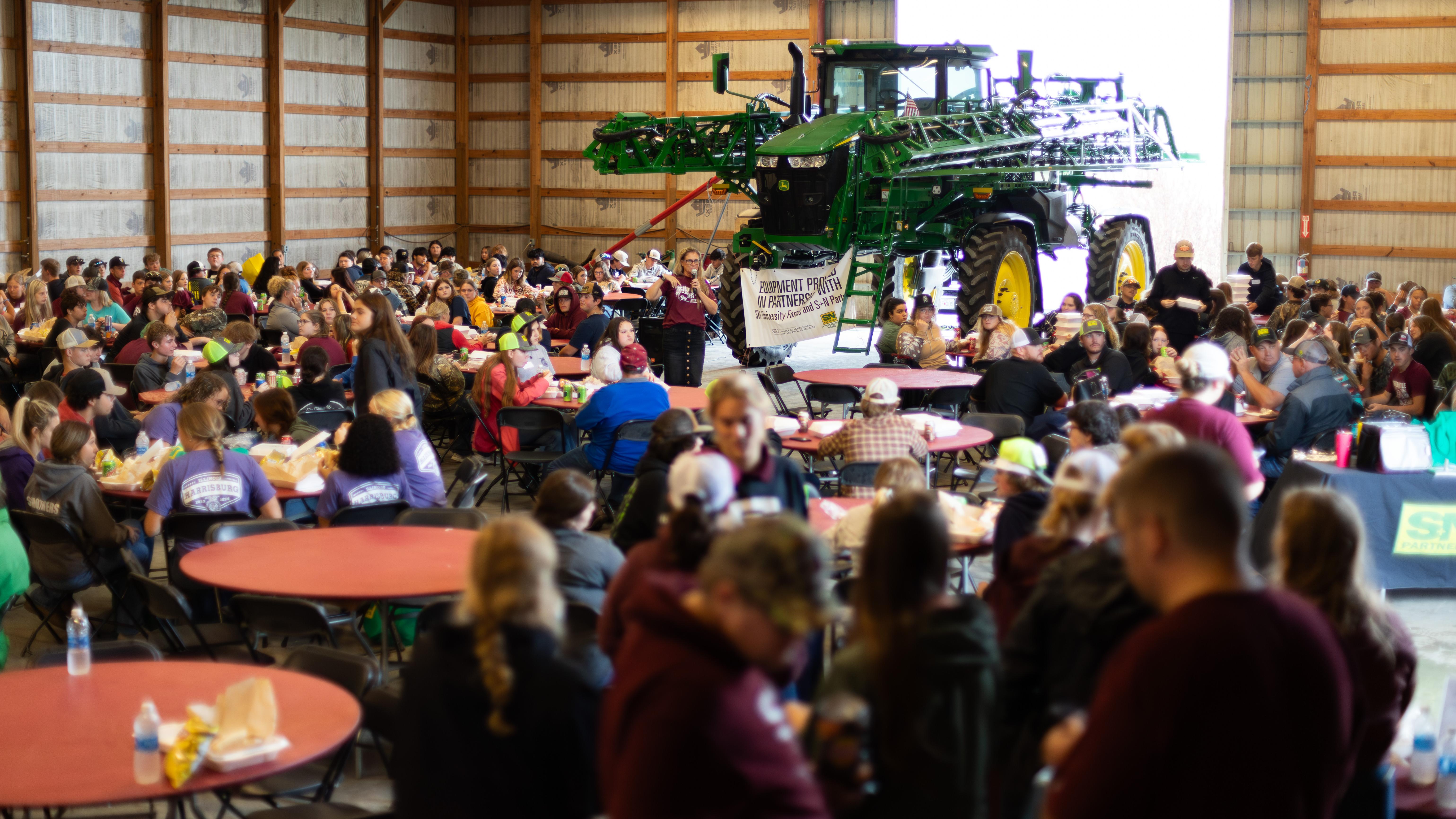 A large group of people, seated around tables. A farm sprayer is seen in the background.