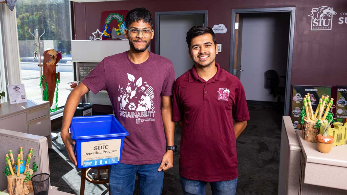 Two men are standing in an office. Both are wearing maroon shirts. One is holding a blue recycling bin.