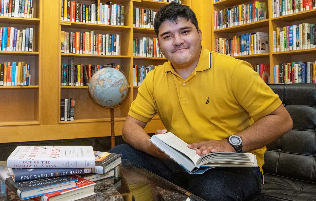 A young man is seated and holding a book. Behind him are shelves of books.