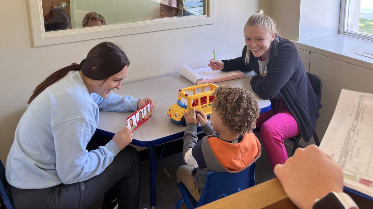 Two young women work with a small child at a table. The child is playing with a toy school bus. One girl is interacting with the child while the other takes notes.