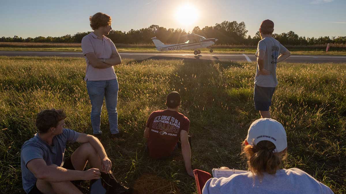 A group of people are standing or seated in the grass as a small airplane takes off.