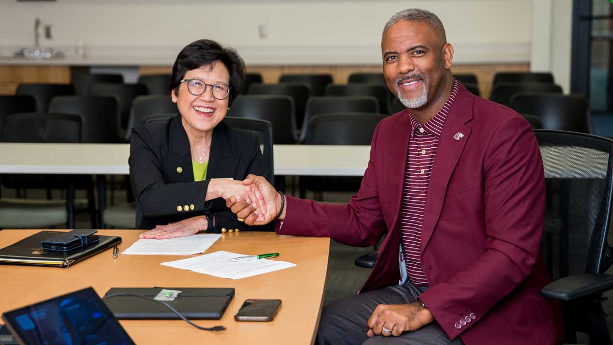 An Asian woman and a black man are seated at a table. They are shaking hands. She is wearing a navy jacket, green shirt, and glasses. He is wearing gray pants, a maroon and white striped shirt, and a maroon jacket.