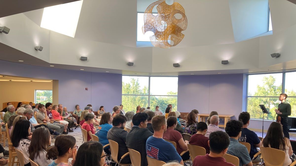 A large group of people, seated, listening to a man speaking and holding a violin.