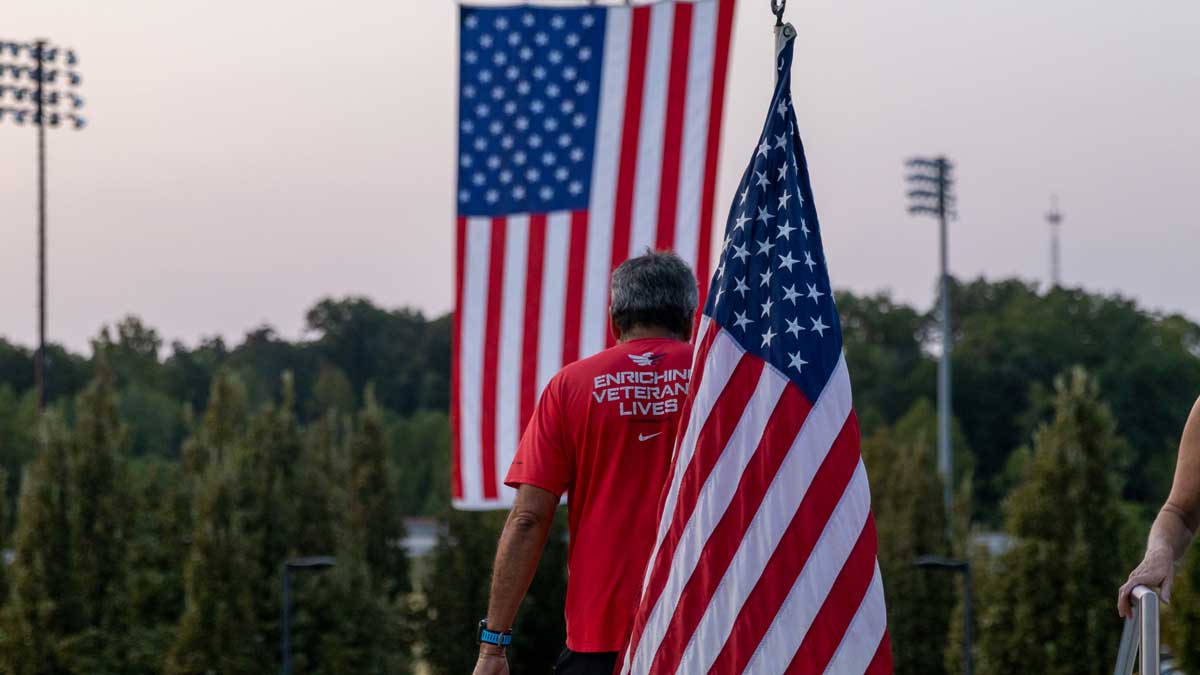 A man is seen from behind while carrying an American flag. There is another flag visible in front of him. He is wearing a red shirt that says Enriching Veterans Lives