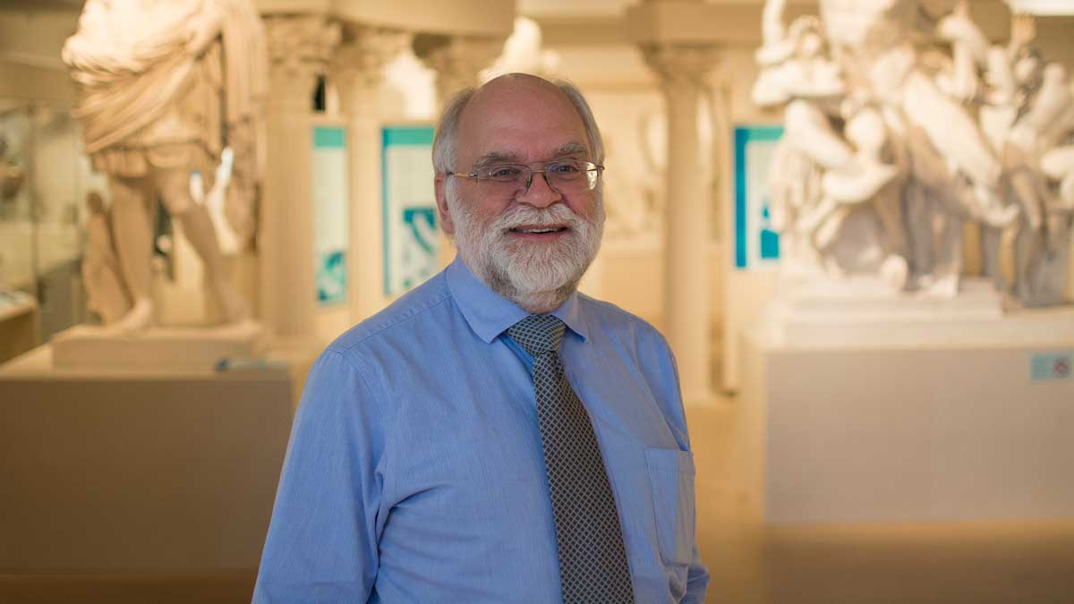 An older man is standing in a large room, smiling for the camera. He has a white beard and is wearing a blue shirt and tie.