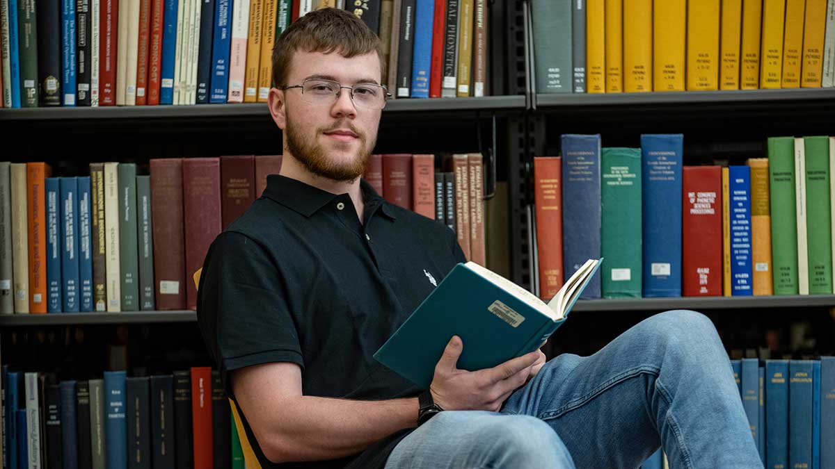 A young man is seated in front of a row of books in a library.