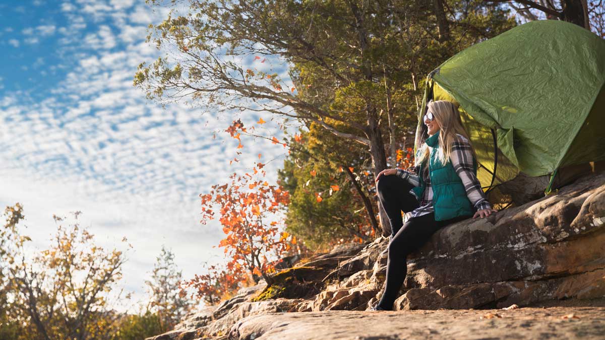 A woman is seated on a rocky bluff beside a tent.