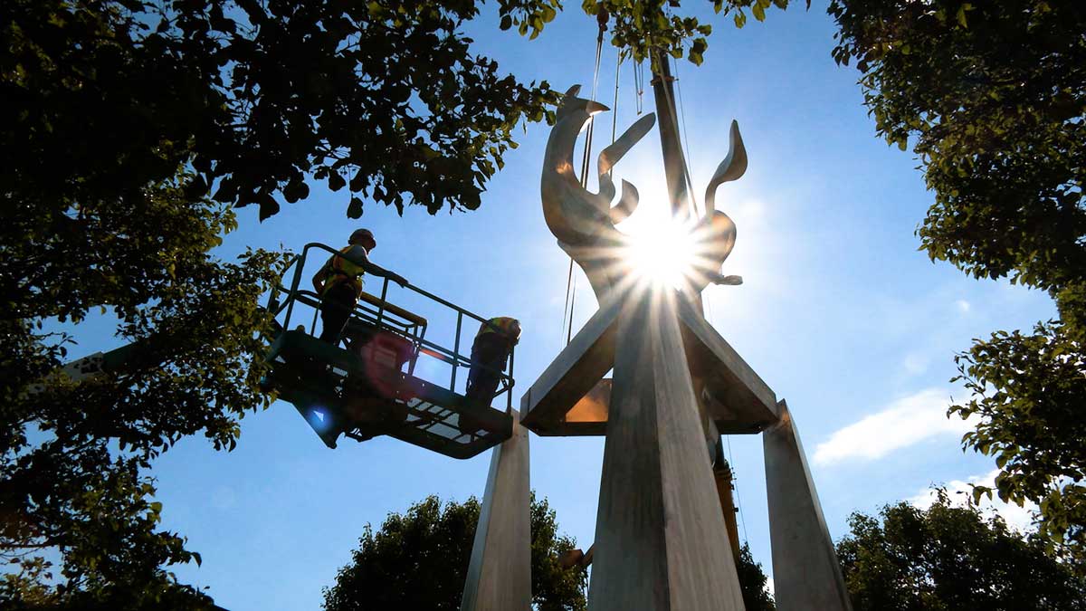 Workers install an art sculpture. A sunburst is visible in the background.