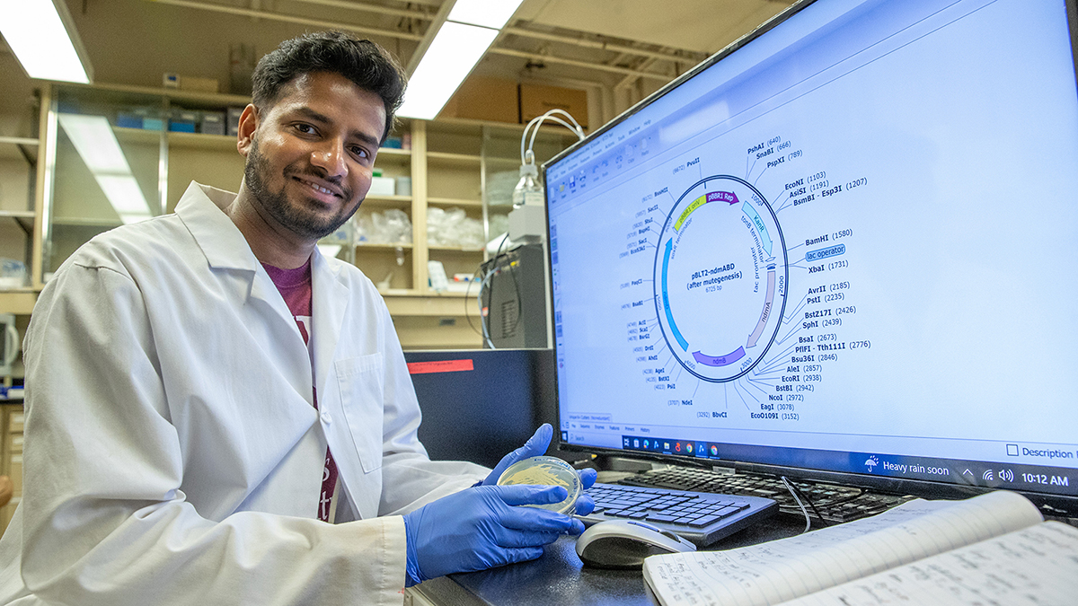 A man seated in front of a computer screen while holding a petri dish.