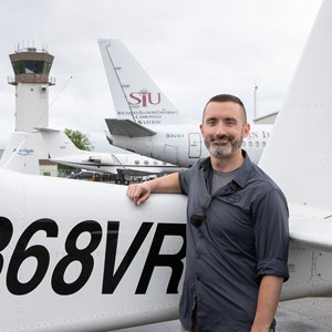 A man is leaning against the tail of an airplane. A air traffic control tower is visible in the background.