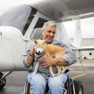 A man seated in a wheelchair, holding a dog in his lap. An airplane is behind him.